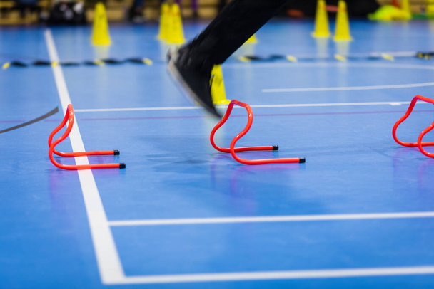 Urheilija hyppää punaisten harjoitusesteiden yli sisätiloissa. Taustalla Futsal-koulutuslaitteet. Jalkapallon esteet, käpyjä ja tikkaat koulutuskentällä - Valokuva, kuva