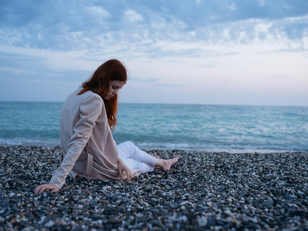 Путешественник с рыжими волосами сидит на пляже и смотрит на океан белой пеной прозрачной воды - Фото, изображение