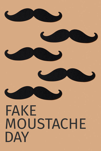 some moustaches, made with cutouts of a black cardboard, and the text fake moustache day on a brown background - Photo, Image