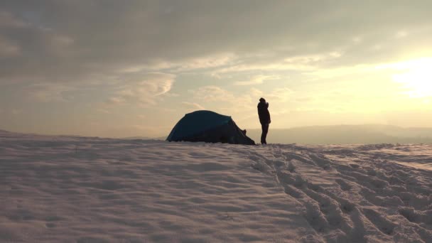 Hiker, happy, enjoy a cup of hot tea from thermos after a day hike, in front of tent, at sunset, in snowy winter mountains - Footage, Video