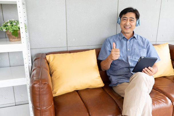 muotokuva peukalot post vanhempi aasialainen mies yllään kuulokkeet ja tilalla tabletti sohvalla - Valokuva, kuva