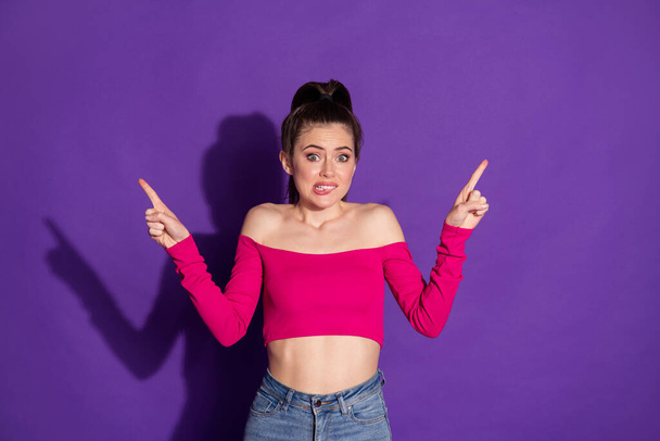 Photo of uncertain young lady raise arms pointing fingers chew lip wear jeans pink top isolated purple color background - Foto, Bild