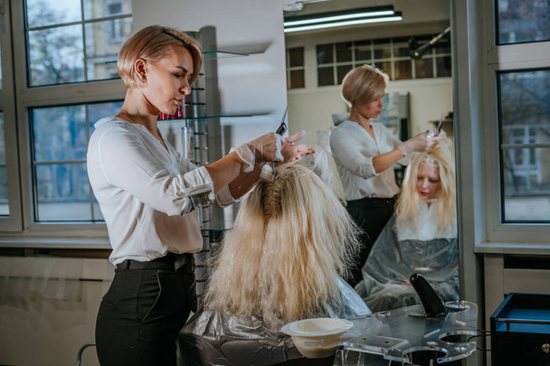 Coiffeur professionnel teinture cheveux blonds de cliente au salon de beauté devant un miroir - Photo, image