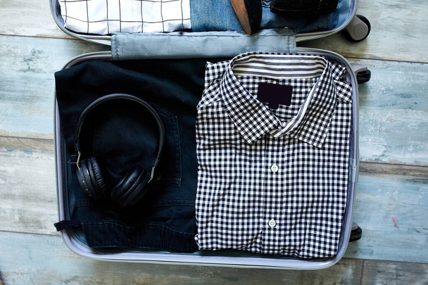 Упаковка чемодан дома с обычными мужскими предметами, вещи - джинсы, рубашки, наушники в чемодане для путешествий, путешествия, отпуска. Копирование пространства. - Фото, изображение