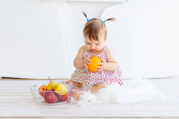 una niña está sentada en una habitación luminosa con un plato de fruta y comiendo una naranja - Foto, imagen
