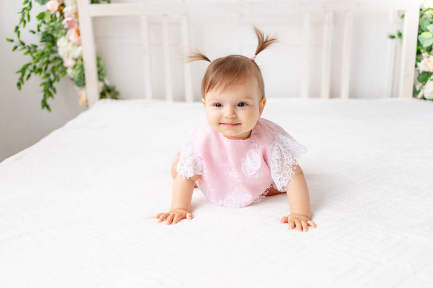 niña alegre de seis meses sentada en una hermosa habitación brillante en una cama blanca en un traje de encaje y sonriendo - Foto, imagen