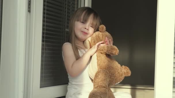 Un gamin insouciant embrasse son jouet. L'enfant joue avec un jouet près de la fenêtre sur le rebord de la fenêtre. Petite fille jouant avec un ours en peluche regardant par la fenêtre. Bonne enfance. Fille joue - Séquence, vidéo