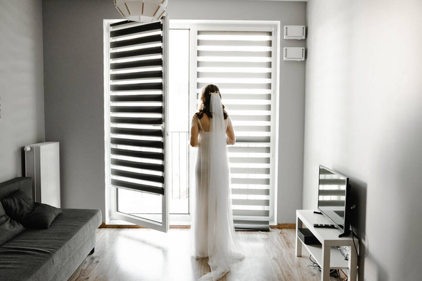 the bride is waiting for the groom in a room with blinds hanging on the window. girl at the window. silhouette of the bride. shutters on the windows. wedding day fees - Valokuva, kuva
