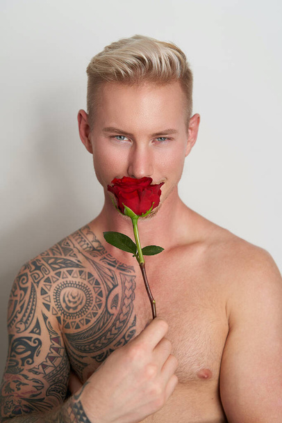 Άντρας εραστής κρύβεται πίσω από ένα λουλούδι με ένα χαμόγελο / Όμορφος νεαρός άνδρας κρατώντας ένα κόκκινο τριαντάφυλλο μπροστά από χαμογελαστό πρόσωπό του - Φωτογραφία, εικόνα