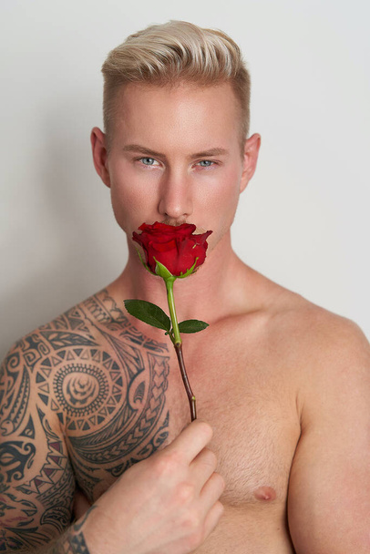 Bir çiçeğin arkasına saklanan erkek aşık. Yüzünün önünde kırmızı bir gül tutan yakışıklı genç adam. - Fotoğraf, Görsel