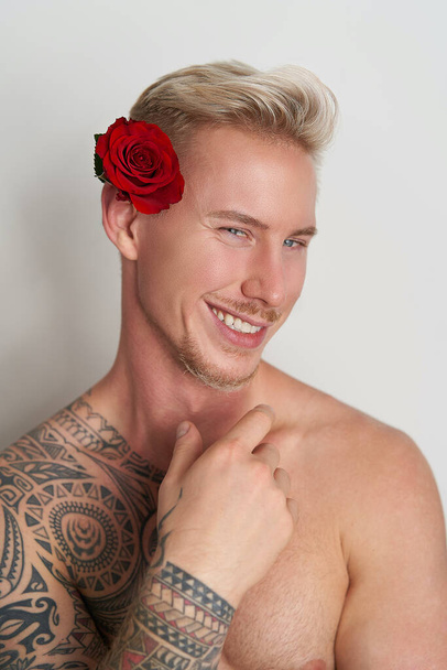 Amante masculino sonriendo con una flor en la oreja / Joven guapo decorado con una rosa roja sonriendo en seductora expectativa - Foto, Imagen