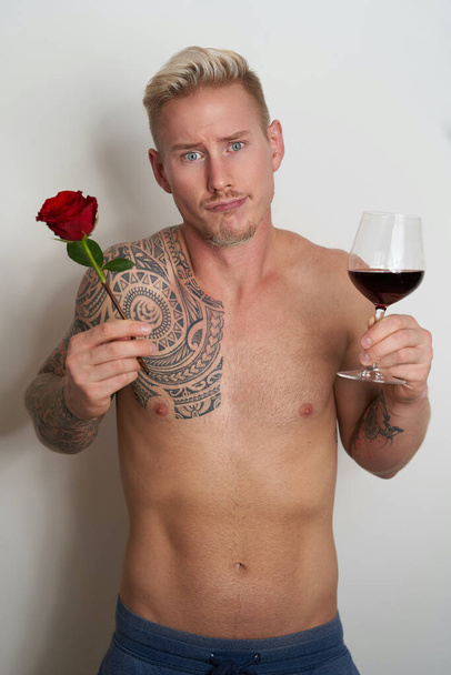 Erkek aşık bir çiçek ve bir bardak şarapla poz veriyor. Yakışıklı genç adam elinde kırmızı bir gül ve içecek tutarken hayal kırıklığına uğramış görünüyor. - Fotoğraf, Görsel