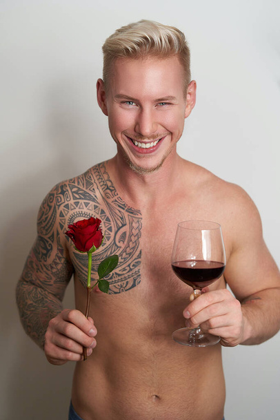 Bir çiçek ve bir bardak şarapla gülümseyen erkek aşık. Kırmızı bir gül ve içecekle sırıtan yakışıklı genç adam. - Fotoğraf, Görsel