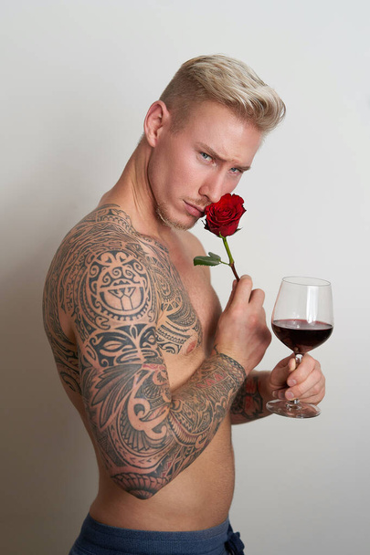 Erkek aşık, bir çiçek ve bir bardak şarapla yan yana poz veriyor. Yakışıklı genç adam, kırmızı bir gül ve içecekle şehvetle poz veriyor. - Fotoğraf, Görsel