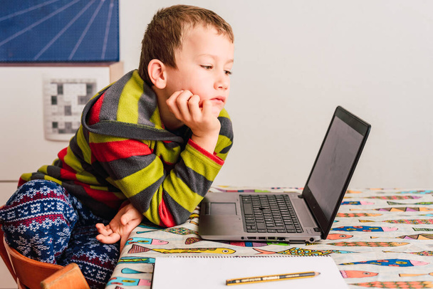 Το αγόρι παρακολουθεί τα μαθήματά του μέσα από έναν υπολογιστή από το σπίτι με μια βαρετή και κουρασμένη χειρονομία. - Φωτογραφία, εικόνα