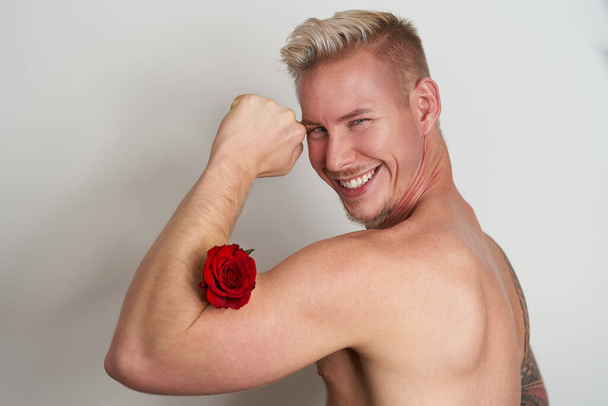 Maschio amante sorridente flessione muscolare con un fiore / Bello giovane uomo sorridente spremendo una rosa rossa nei suoi bicipiti - Foto, immagini
