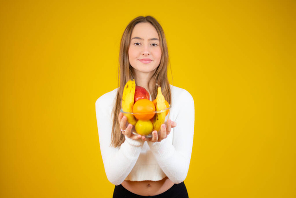 Happy beautiful girl holding bowl of fresh fruits, studio shot, isolated over yellow background - Photo, Image