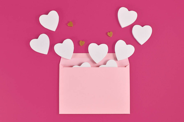 White and golden wooden hearts spilling out of pastel pink envelope on pink background - Foto, afbeelding