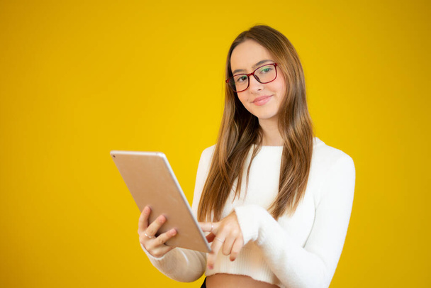 Photo of happy young girl standing isolated over yellow wall background. Looking aside using tablet computer. - Φωτογραφία, εικόνα