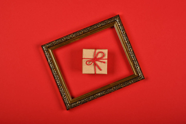 Close up one brown paper giftbox in golden frame over red background, flat lay, elevated top view, directly above - Fotografie, Obrázek