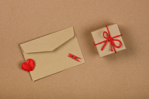 Close up Valentine gift box with red twine bow and card greeting envelope with wooden clothespins and hearts over brown paper background, flat lay, elevated top view, directly above - Фото, зображення