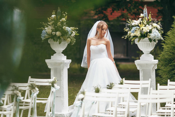 Schöne Braut in einem prächtigen Brautkleid posiert inmitten von Grün auf der Straße. Mädchen posiert in einem Hochzeitskleid für Werbung. Brautkonzept für Werbekleider. - Foto, Bild