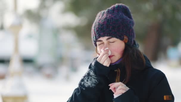Winter koud en loopneus. Portret van een jonge vrouw staat op straat en blaast haar neus met een papieren zakdoek. Met sneeuw bedekte straat op de achtergrond. - Video