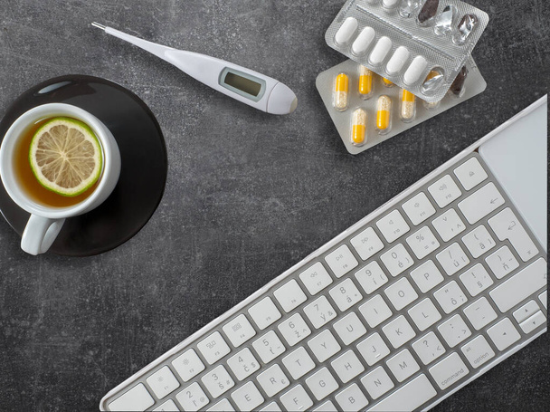 Witte computer toetsenbord, zwarte kop thee, digitale thermometer en blaren van pillen op beton werktafel. Overheadshot met kopieerruimte. - Foto, afbeelding