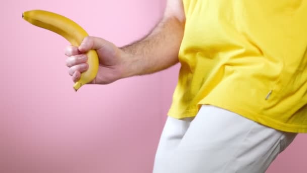 Erősség. Az ember egy banánt tart a kezében a nemi szervek szintjén, úgy játszik vele, mint egy pisztollyal. Közelről. Rózsaszín háttér. A férfiak egészségének fogalma. - Felvétel, videó