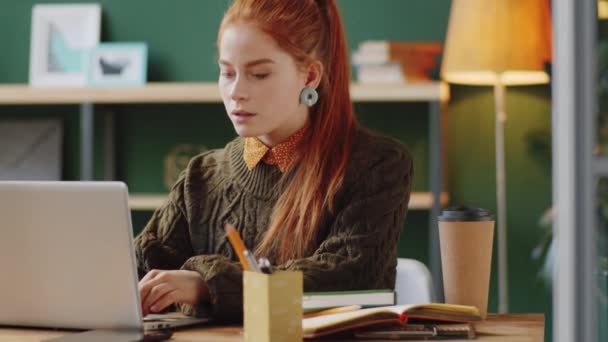 Giovane donna d'affari rossa che lavora su laptop e prende appunti alla scrivania in un ufficio moderno con pareti verde scuro - Filmati, video