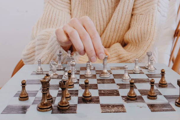 Frau im beigen Pullover sitzt vor Schachbrett und plant Strategie. Handgesten der Unsicherheit. Mädchen spielen Schach in Innenräumen und Denkposition, finden Gewinnzug, Strategie. Hobby-Konzept - Foto, Bild
