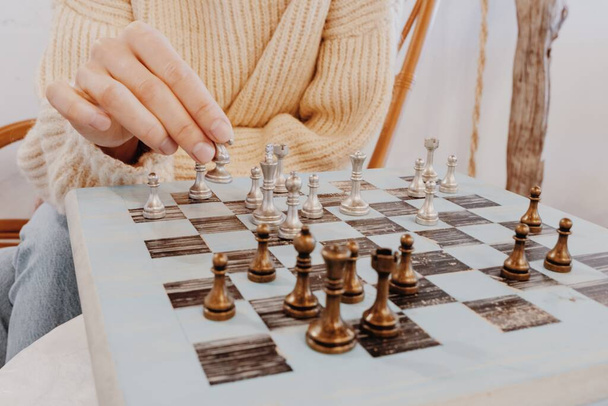 Woman in beige sweater sitting in front of chess board and planning strategy. hand gestures of uncertainty. Girl playing chess indoor and thinking position, find winning move, strategy. Hobby concept - Photo, Image