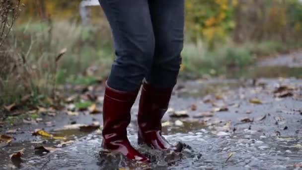Primer plano de una mujer con botas de goma roja saltando en un charco fangoso de otoño después de la lluvia - Imágenes, Vídeo
