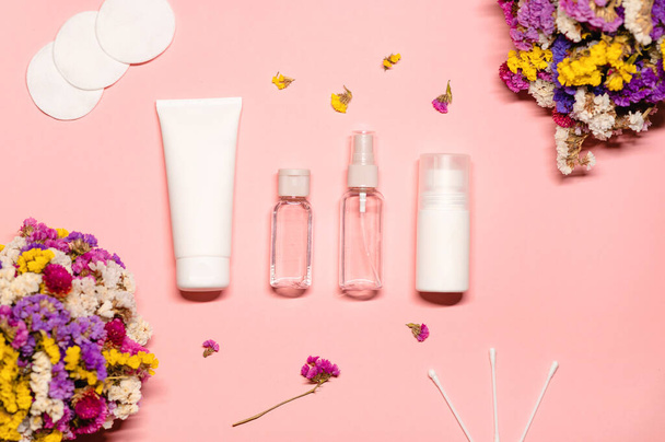 Flat lay foto com produtos de tratamento de pele: creme facial, soro, toner, limpador, almofadas de algodão e botões com flores vivas em um fundo rosa. - Foto, Imagem