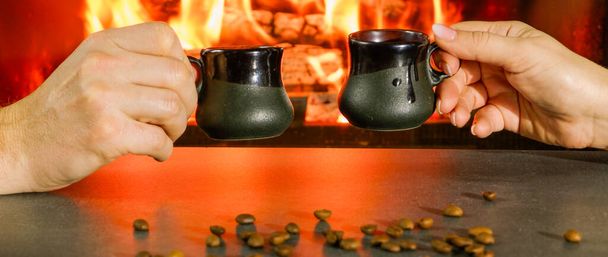 Romantic evening. Couple having a cup of coffee near the fire. Close-up of hands in the backlight of a burning fireplace. - 写真・画像