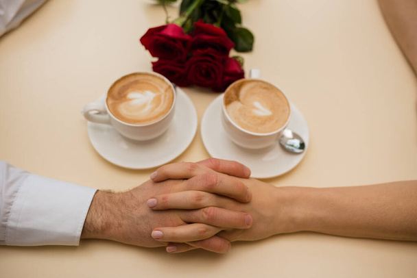 manos unidas en una mesa con dos tazas de capuchino con un ramo de rosas rojas - Foto, imagen