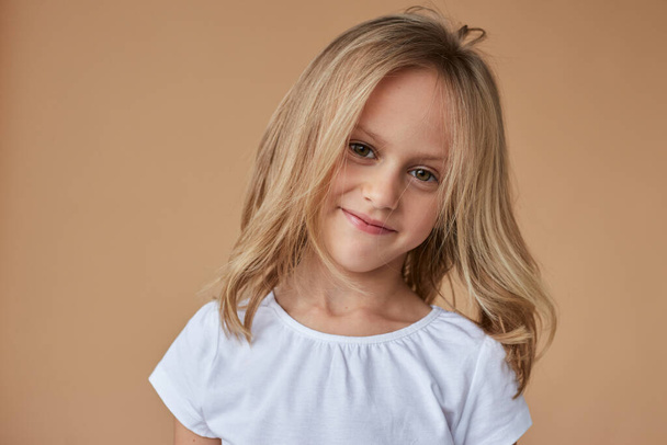 Nahaufnahme Porträt des hübschen kleinen Mädchens mit welligen blonden Haaren, gekleidet in weiße Kleidung, über beigem Hintergrund. - Foto, Bild