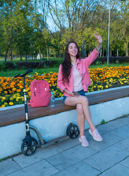 Woman in summer in a city park waves her hand, happy smiles rejoices, meeting and a date, scooter shop bag and bottle with water. Beautiful girl on vacation. - Фото, зображення