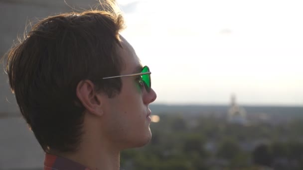 Profilja fiatalember nézi panoráma a város tetőről, és egyenesíti a hajat. Jóképű fickó napszemüvegben a tetőről nézi a városképet, és élvezi a gyönyörű kilátást. Oldalnézet Bezárás Lassú mozgás - Felvétel, videó