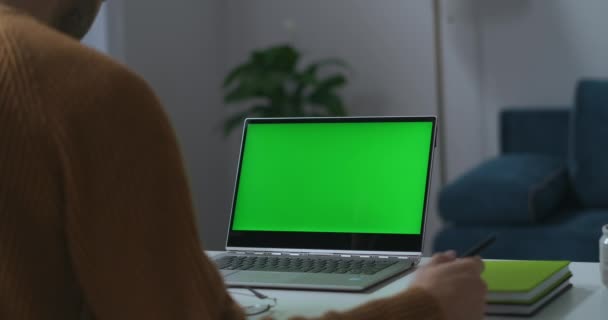 laptop zöld képernyő a munkaasztalon otthoni irodában, kilátás vállán keresztül férfi, távoli munka és szabadúszó - Felvétel, videó