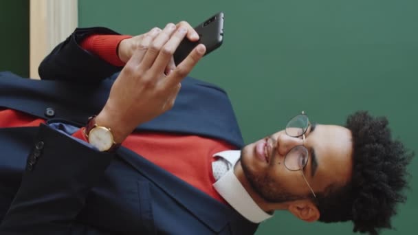 Вертикальный крупный план стильного бизнесмена смешанной расы, стоящего напротив зеленой стены и обмена сообщениями на смартфоне - Кадры, видео