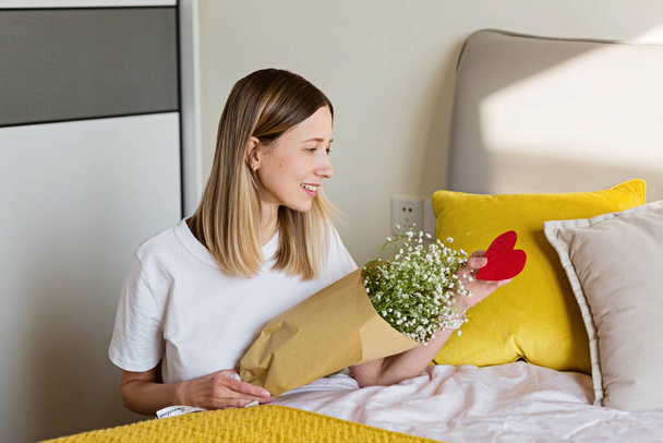 Mooie jonge Kaukasische vrouw met boeket bloemen in de slaapkamer. Fijne Valentijnsdag. verre relatie en liefde concept - Foto, afbeelding