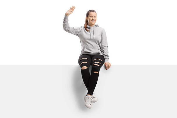 Cheerful young female in a hoodie and ripped trousers sitting on a panel and waving isolated on white background - Foto, immagini