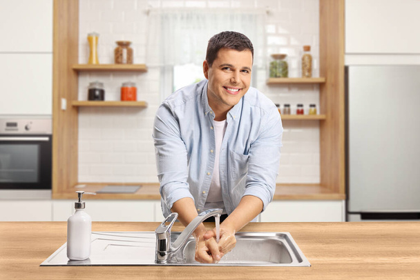 Young man washing hands in a kitchen at home and smiling at camera - Valokuva, kuva