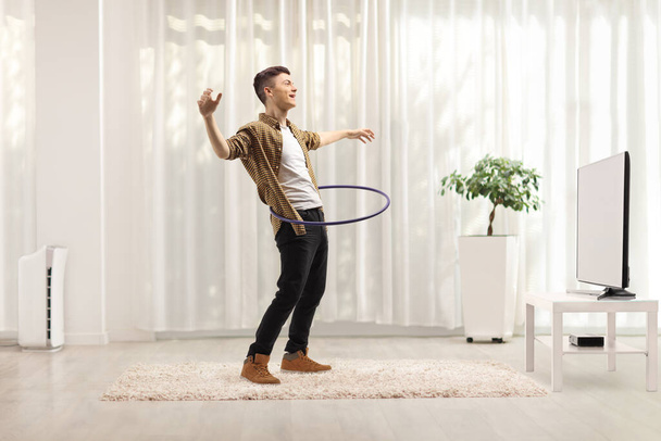 Full length shot of a young man spinning a hula hoop at home in front of tv - Fotó, kép