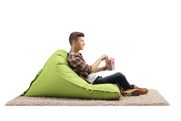 Guy sitting on a bean bag armchair with a box of popcorn isolated on white background - Φωτογραφία, εικόνα