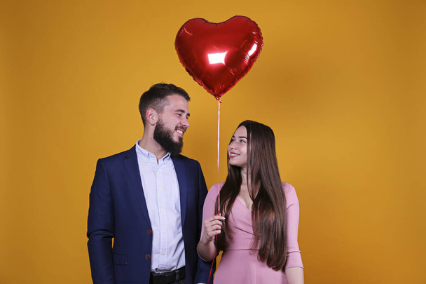 Happy Valentine's day concept. Studio shot of couple in love holding a heart shaped balloon. Portrait of bearded guy and girl in pink dress posing over yellow wall background. Copy space, casual style - Foto, imagen