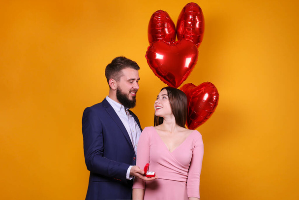 Bearded man proposing to his beautiful girlfriend on st. Valentine's day. Guy with perfectly groomed facial hair and young woman in low-cut dress getting engaged. Yellow wall background, copy space. - Photo, image