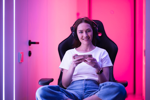 Young girl playing mobile online game on a smart phone while sitting on gaming chair in colorful neon light room. - Photo, Image