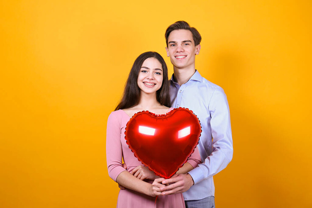 Happy Valentine's day concept. Studio shot of couple in love holding a heart shaped balloon, showing affection. 14th february - the lovers day. Yellow wall background, copy space, front view portrait. - Photo, Image